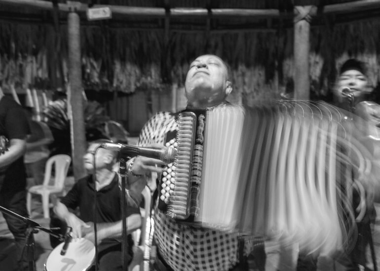A vallenato accordionist plays with his head tilted back and eyes closed, while the accordion bellows blur with movement. Other musicians perform around him in this black-and-white photograph.
